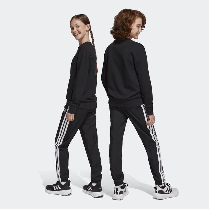 Two children modeling black Adidas Essentials Climacool Tracksuit Bottoms with white stripes, wearing matching black