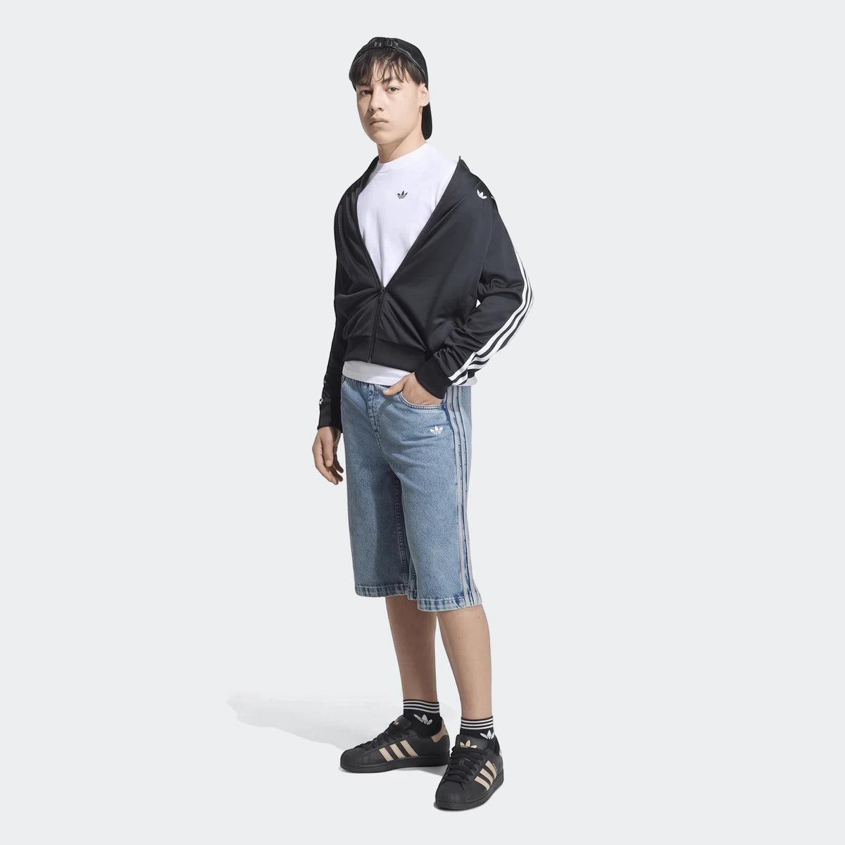 Young man wearing Adidas black track jacket, white t-shirt, blue denim jorts, and black Superstar sneakers, full body shot.
