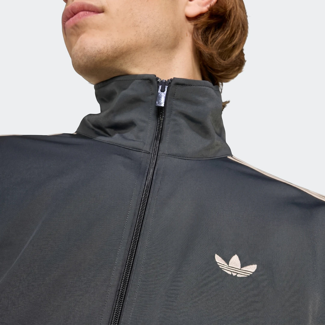 Close-up of a man wearing a black Adidas Firebird Track Top, showing the iconic beige Trefoil logo and zipper detail.