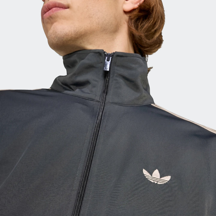Close-up of a man wearing a black Adidas Firebird Track Top, showing the iconic beige Trefoil logo and zipper detail.