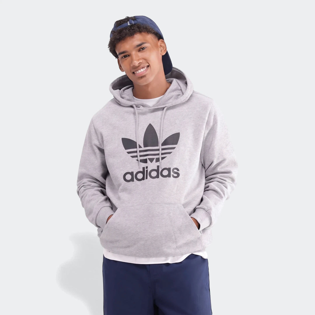 A smiling young man in a grey men's Adidas Trefoil Hoodie and a blue cap, standing with hands in pockets.