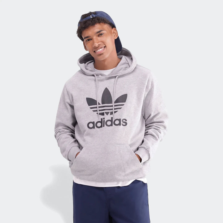 A smiling young man in a grey men's Adidas Trefoil Hoodie and a blue cap, standing with hands in pockets.