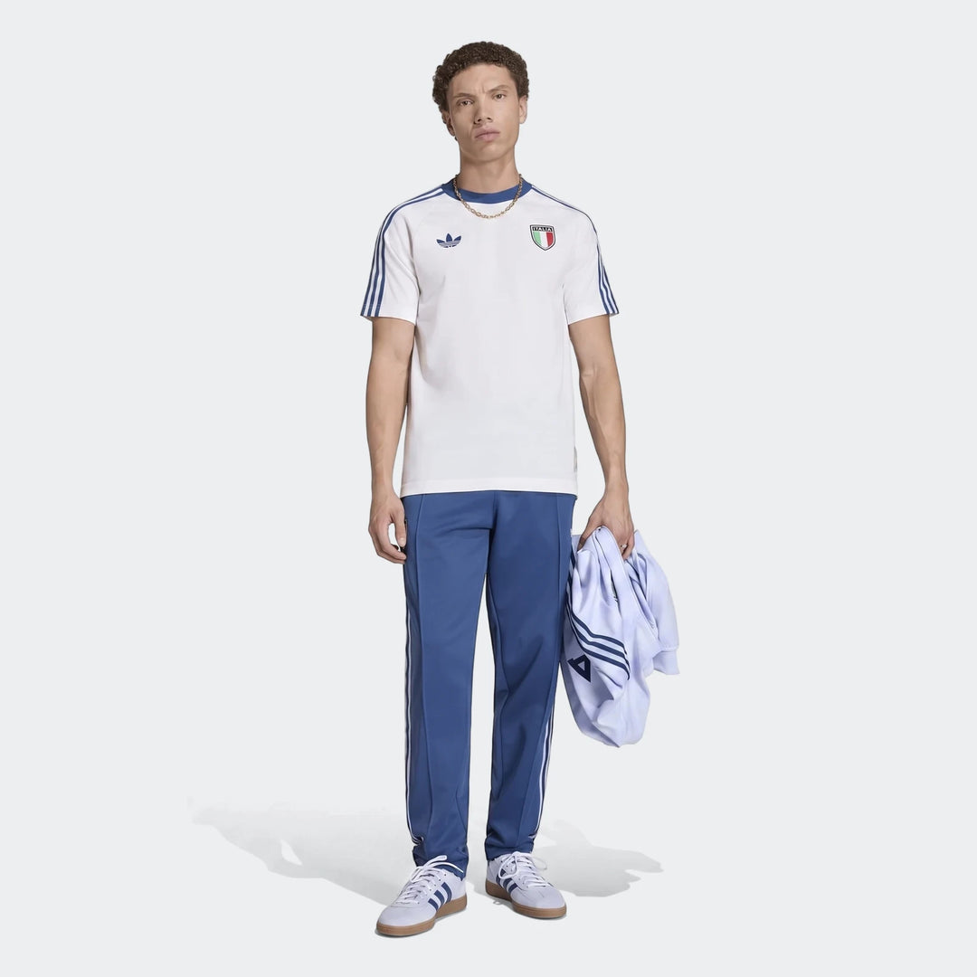 A man models a white Adidas Originals Italy graphic t-shirt with blue stripes, blue track pants, and white sneakers.