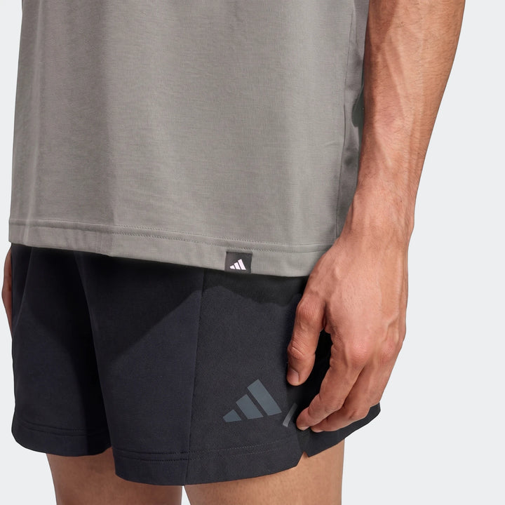 Close-up side view of a male model wearing a grey Adidas Primelift Graphic T-Shirt with a logo tag and black Adidas shorts.