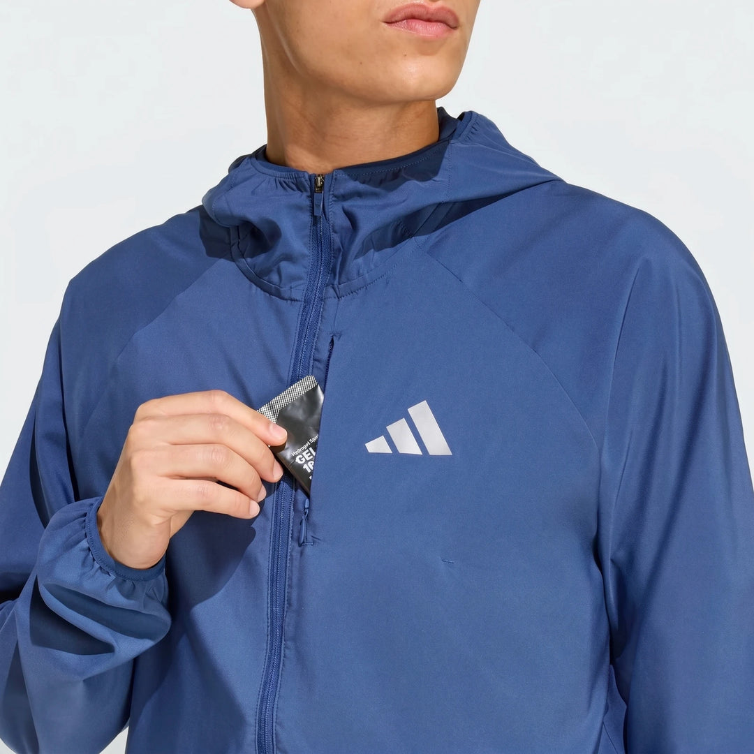 Blue Adidas Run Essentials Jacket with a hood, worn by a person from the waist up, reaching into a chest pocket with a black