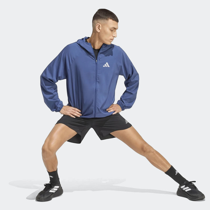 Male model in a blue Adidas Run Essentials Jacket and black shorts, striking a dynamic stretching pose.