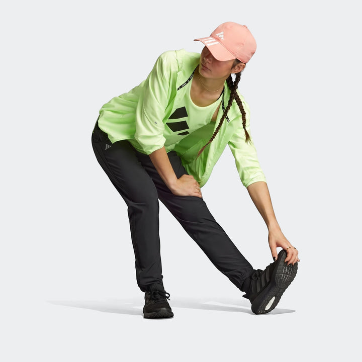 Woman in black Adidas Run It Tko Joggers stretching, wearing a neon yellow-green windbreaker and a pink cap.