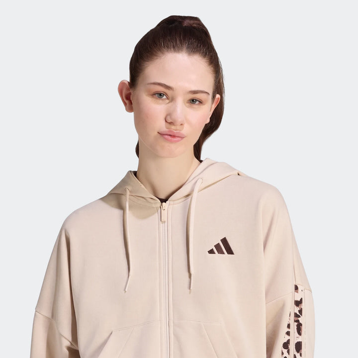 Woman wearing a beige Adidas Seasonal Essentials Animal Print 3-Stripes Full-Zip Hooded Track Top with animal print detail
