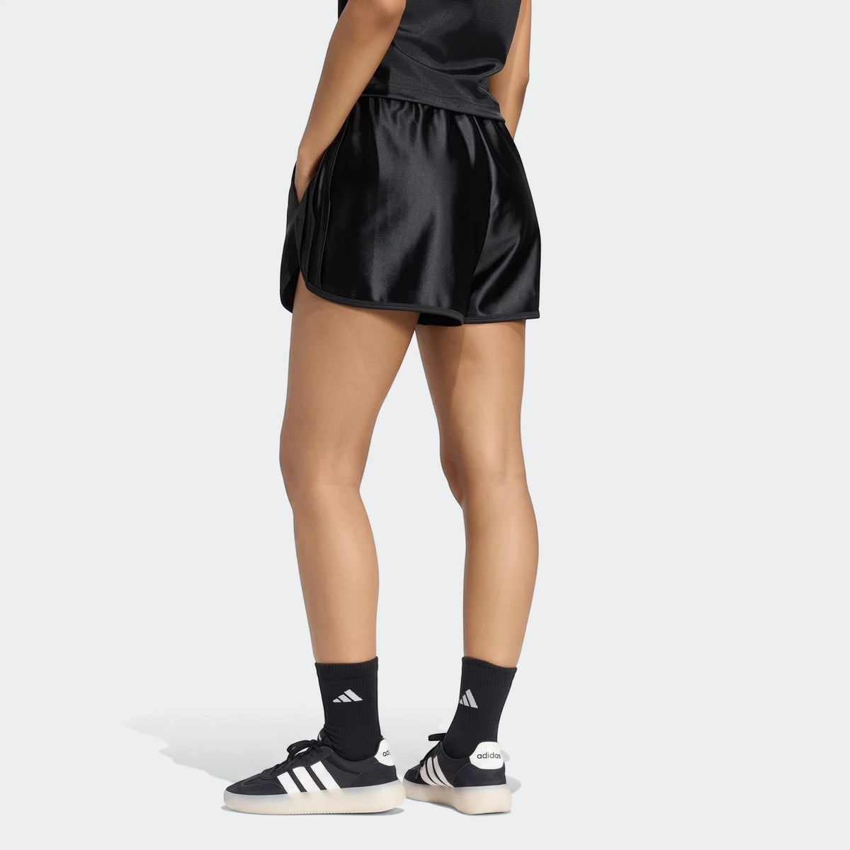 Women's black Adidas Seasonal Essentials satin shorts worn by a model from the back, paired with black socks and sneakers.
