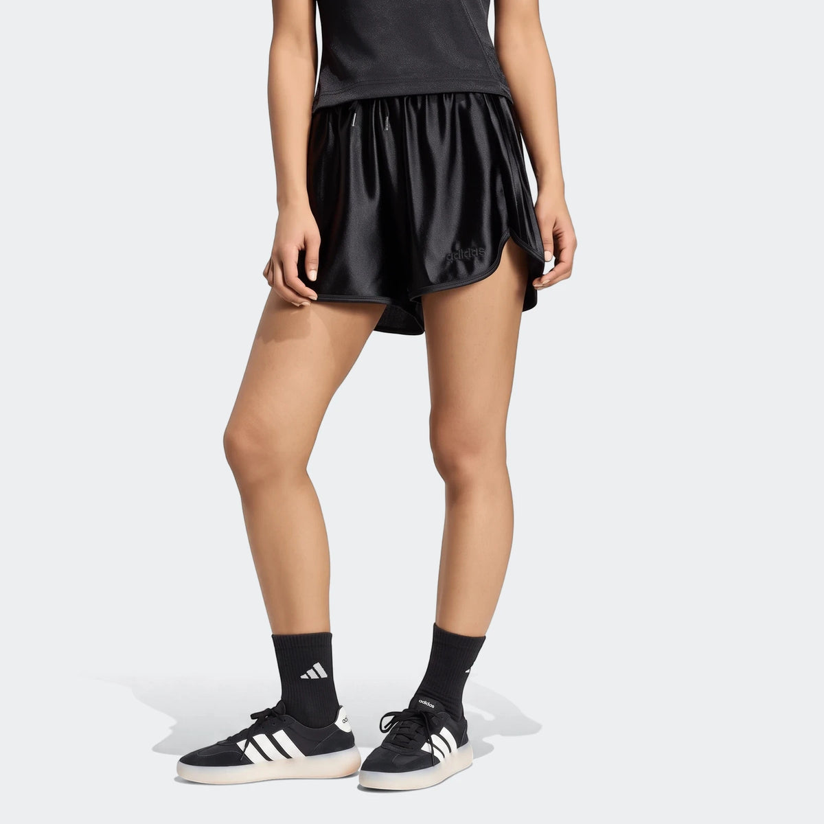 Woman wearing black Adidas Seasonal Essentials satin shorts, black top, black socks, and black and white Adidas sneakers,