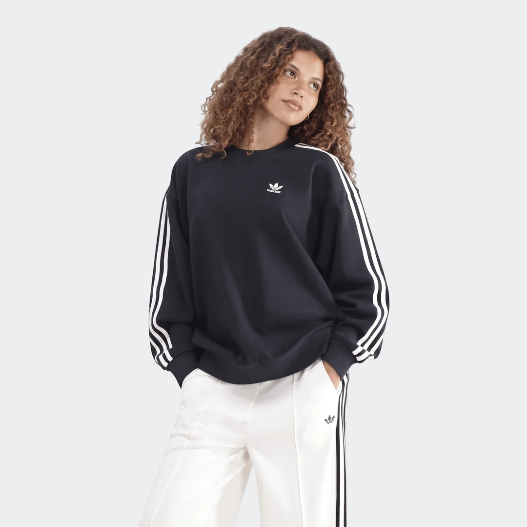 Model wearing a black Adidas Sportswear 3-Stripes Oversized Crew Sweatshirt with white arm stripes and white track pants.