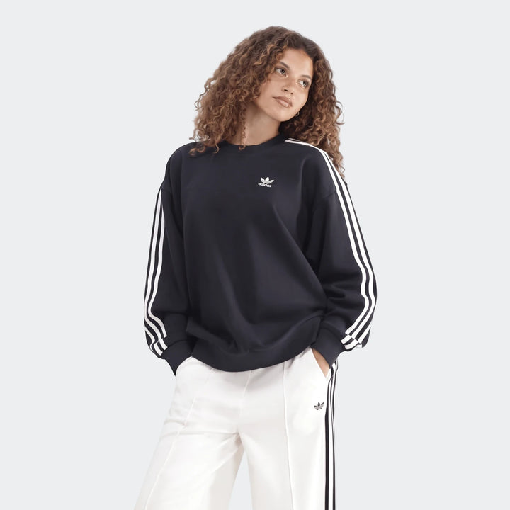 Model wearing a black Adidas Sportswear 3-Stripes Oversized Crew Sweatshirt with white arm stripes and white track pants.