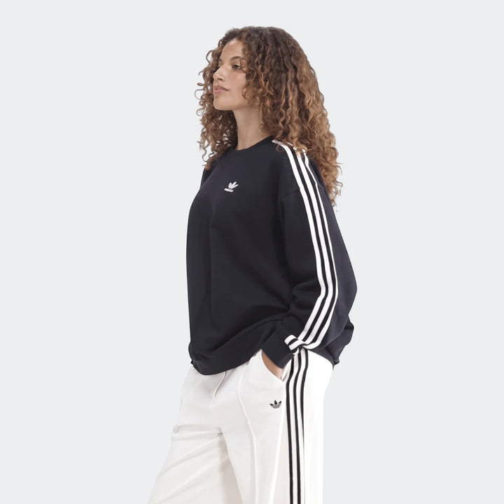 Woman wearing a black Adidas Sportswear 3-Stripes Oversized Crew Sweatshirt and white track pants, posing with hands in