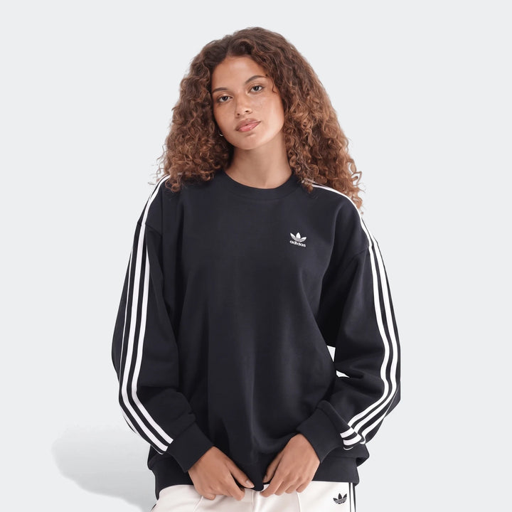 Woman wearing a black Adidas Sportswear 3-Stripes Oversized Crew Sweatshirt and light pants, captured in a front view.
