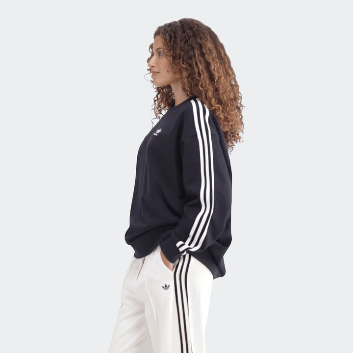 Woman wearing a navy Adidas Sportswear 3-Stripes Oversized Crew Sweatshirt with white stripes and white track pants, profile