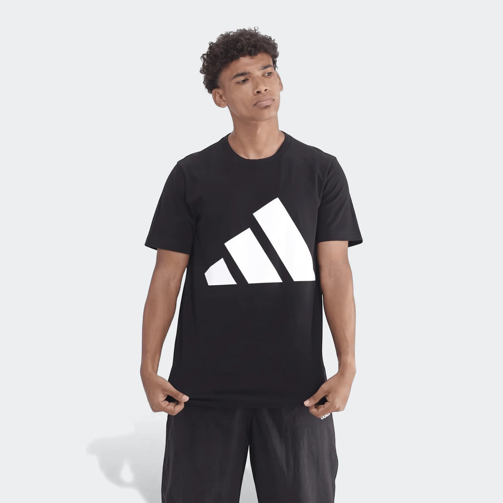 Young man wearing a black Adidas Sportswear Badge Of Sport T-shirt and black shorts, standing on a gray background. The