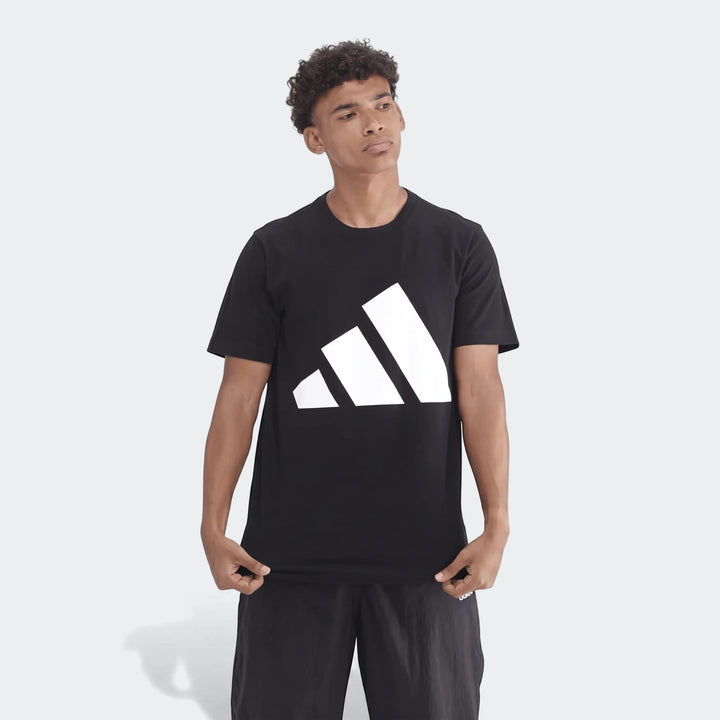 Young man wearing a black Adidas Sportswear Badge Of Sport T-shirt and black shorts, standing on a gray background. The