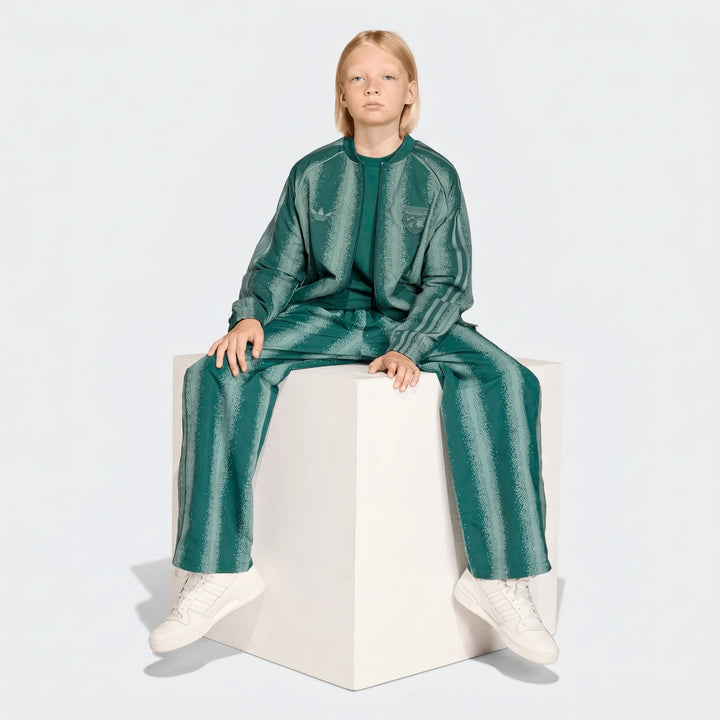 Young person wearing green striped Adidas Sst Loose Track Pants and matching jacket, sitting on a white cube.