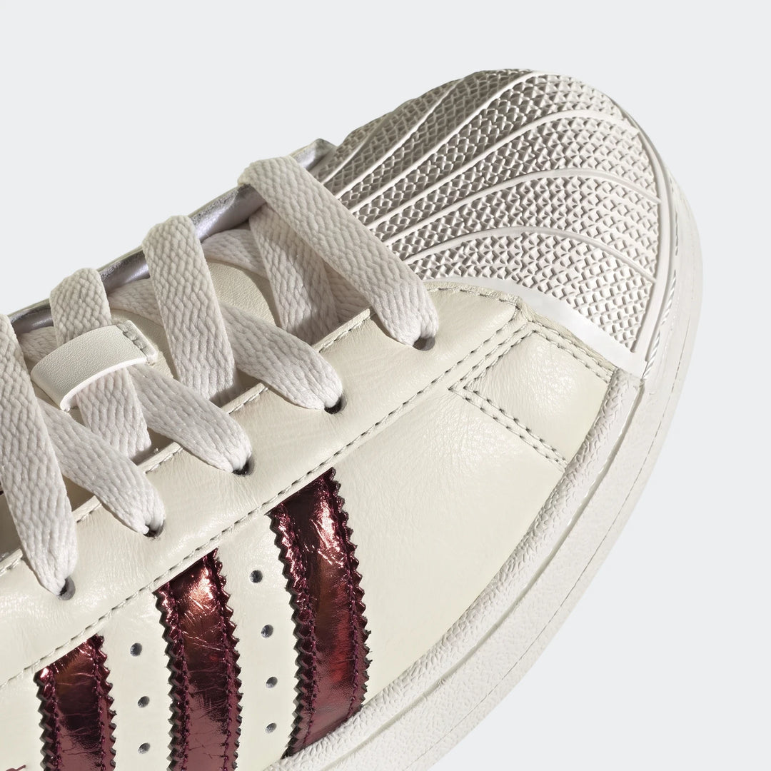 Close-up front view of cream Adidas Superstar II shoes with white laces, textured shell toe, and metallic reddish-bronze
