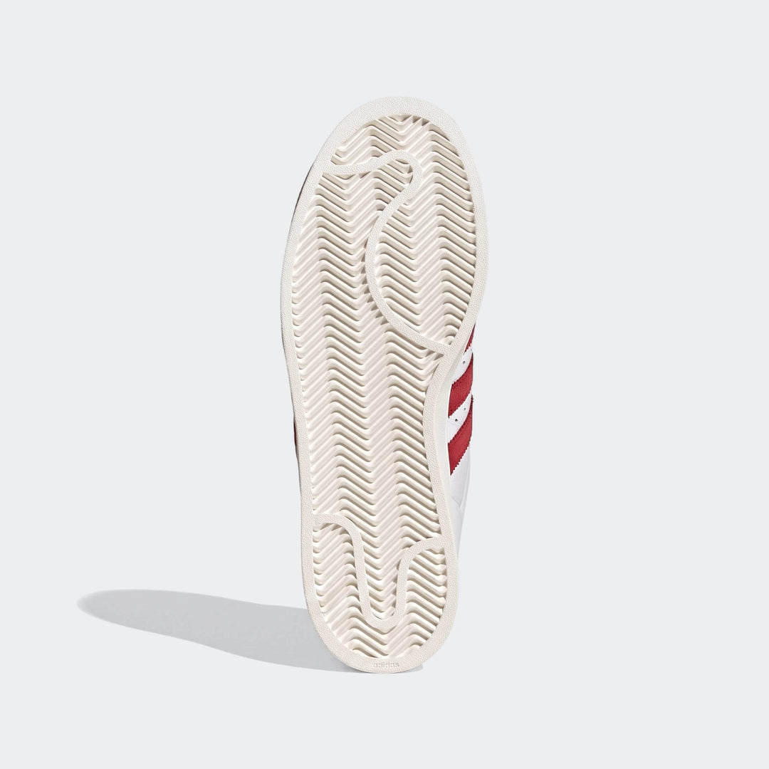 White Adidas Superstar II shoes with red stripes, showcasing the textured outsole pattern from a bottom-up perspective.