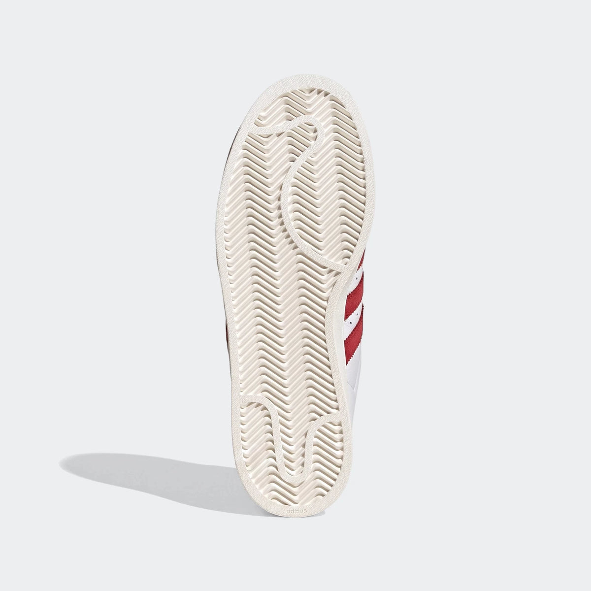 White Adidas Superstar II shoes with red stripes, showcasing the textured outsole pattern from a bottom-up perspective.