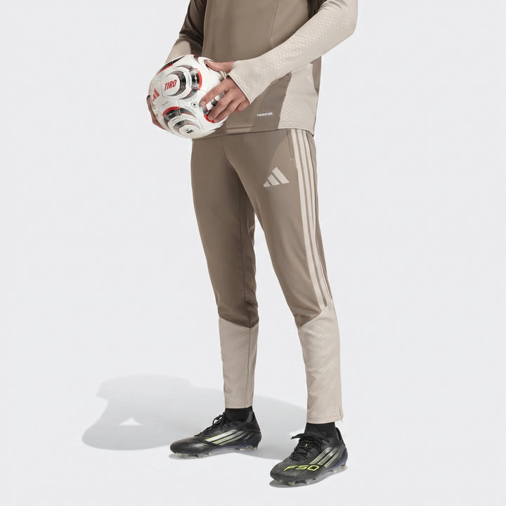 Person wearing Adidas Tior26C brown track pants with white stripes, black soccer cleats, and holding a white soccer ball.