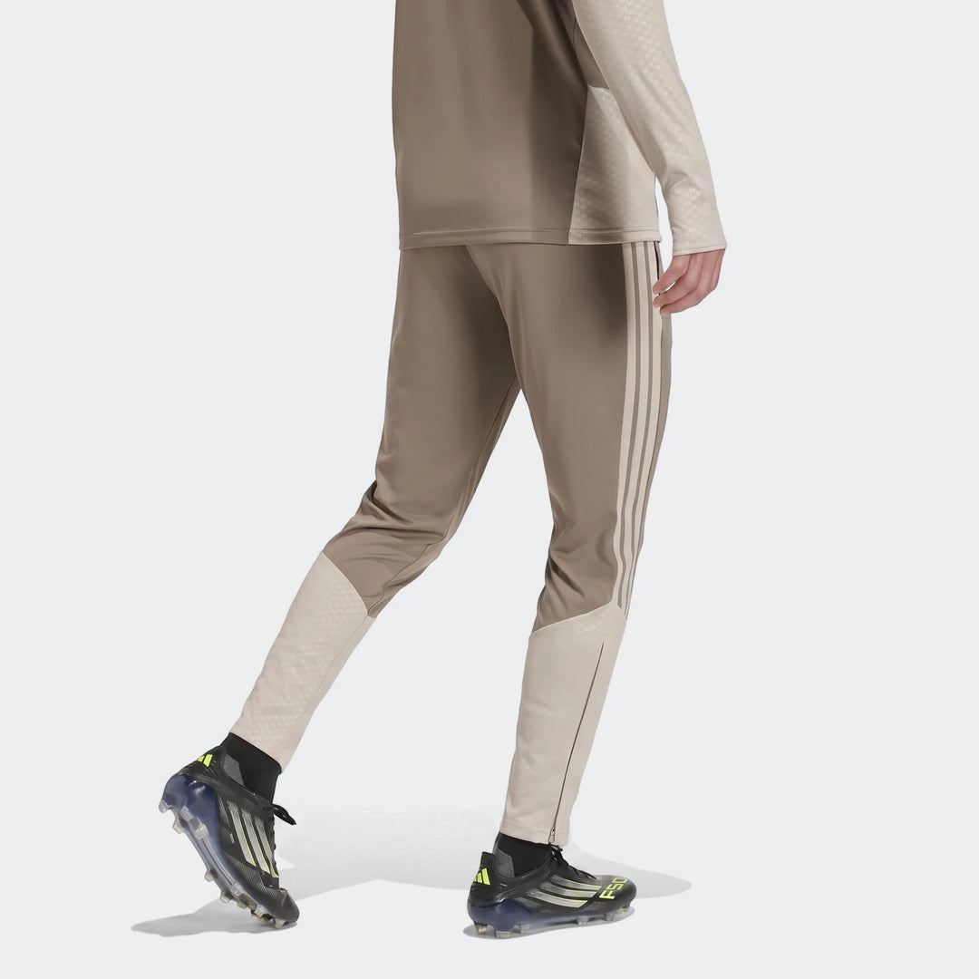 Person wearing Adidas Tior26C brown track pants with light stripes, showing textured panels, an ankle zipper, and black