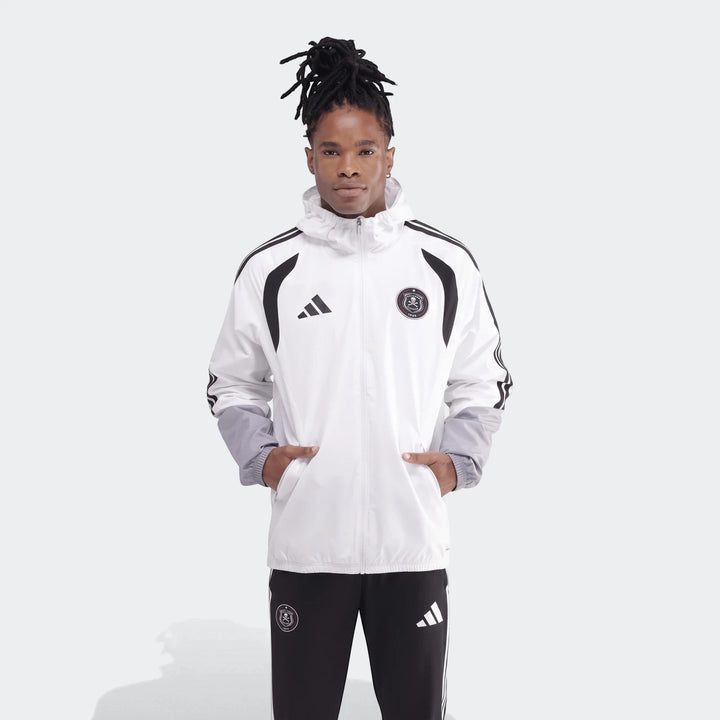 Male model wearing a white and black Adidas Tiro 26C All-Weather Jacket and black pants, standing with hands in pockets.