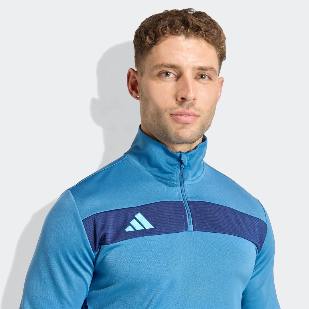 Male model wearing a blue Adidas Tiro Essentials Quarter-Zip Track Top, front view, in a studio setting.