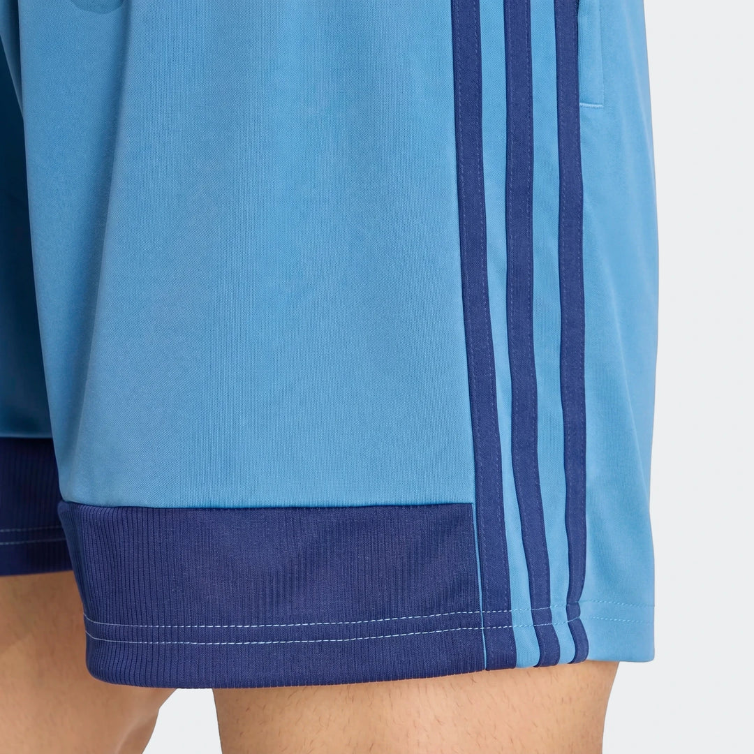Close-up of light blue Adidas Tiro Essentials shorts with dark blue three-stripes and ribbed cuffs, worn on a person.