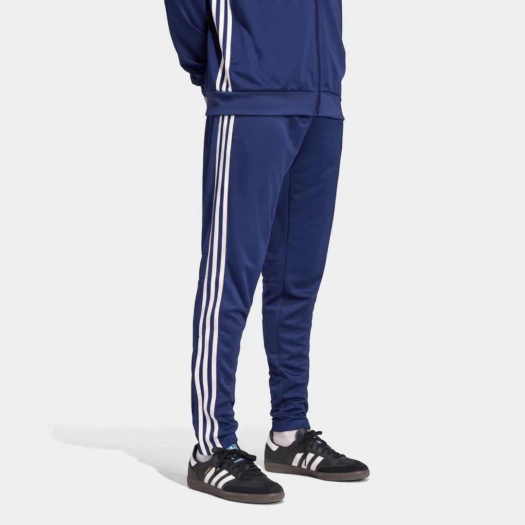 Model wearing navy blue Adidas Tiro Essentials track pants and matching jacket with white three-stripes and black sneakers.