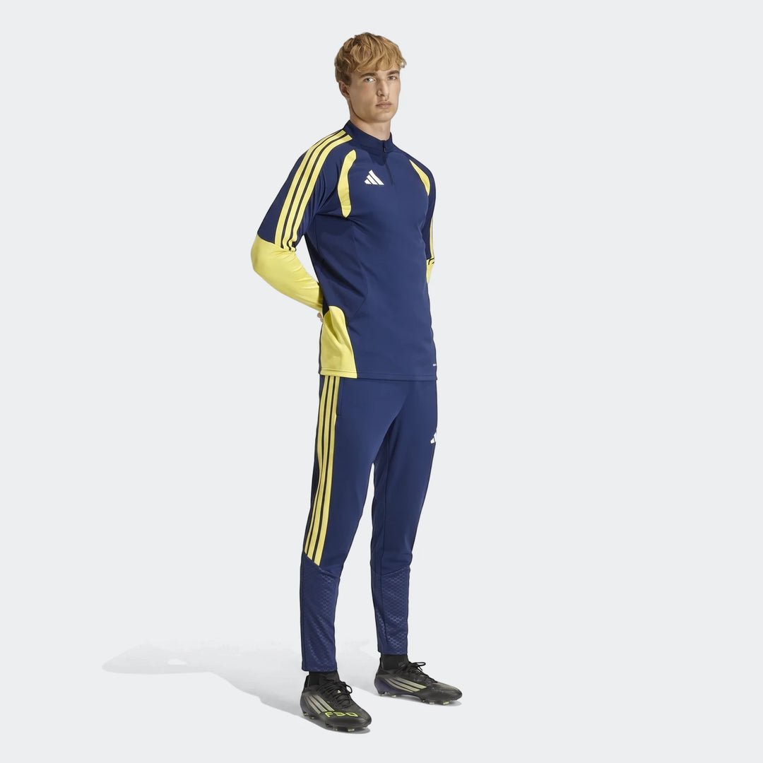 Male model wearing Adidas Tiro26 C Pant and matching long-sleeve top in navy blue and yellow, with black soccer cleats.