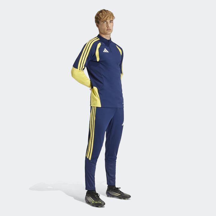Male model wearing Adidas Tiro26 C Pant and matching long-sleeve top in navy blue and yellow, with black soccer cleats.