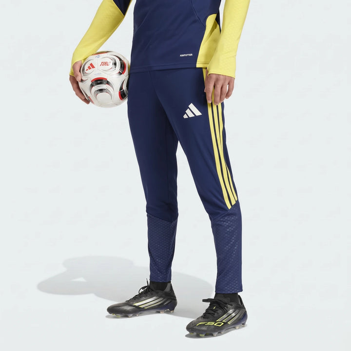 A person wears navy blue Adidas Tiro26 C training pants with yellow stripes and black soccer cleats, holding a white and red