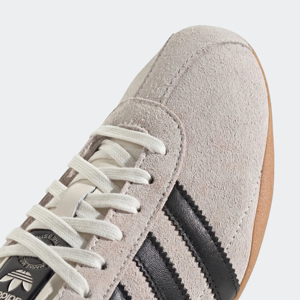 Close-up of an Adidas Tokyo shoe, showcasing the beige suede upper with black serrated three-stripes, cream laces, and a