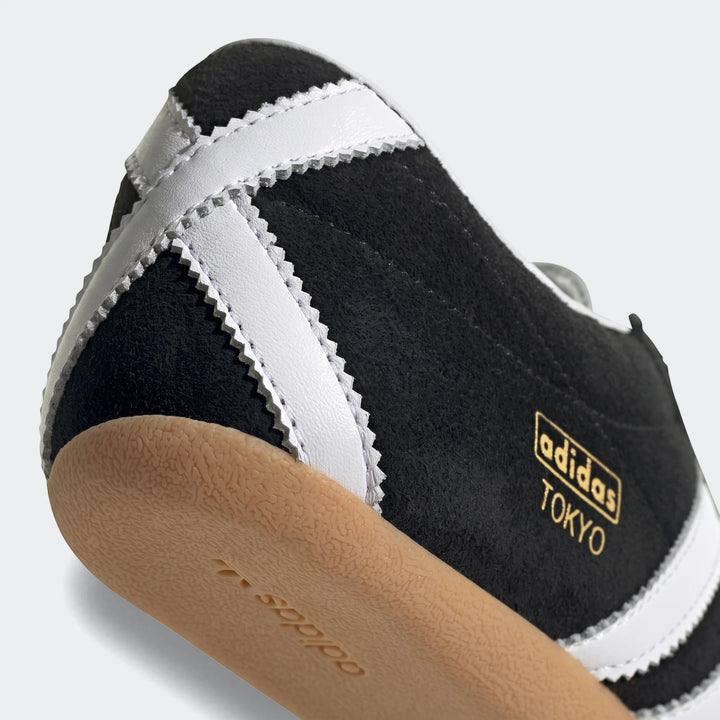 Close-up of a black suede Adidas Tokyo shoe with white serrated stripes and a tan gum sole, viewed from the side and heel.
