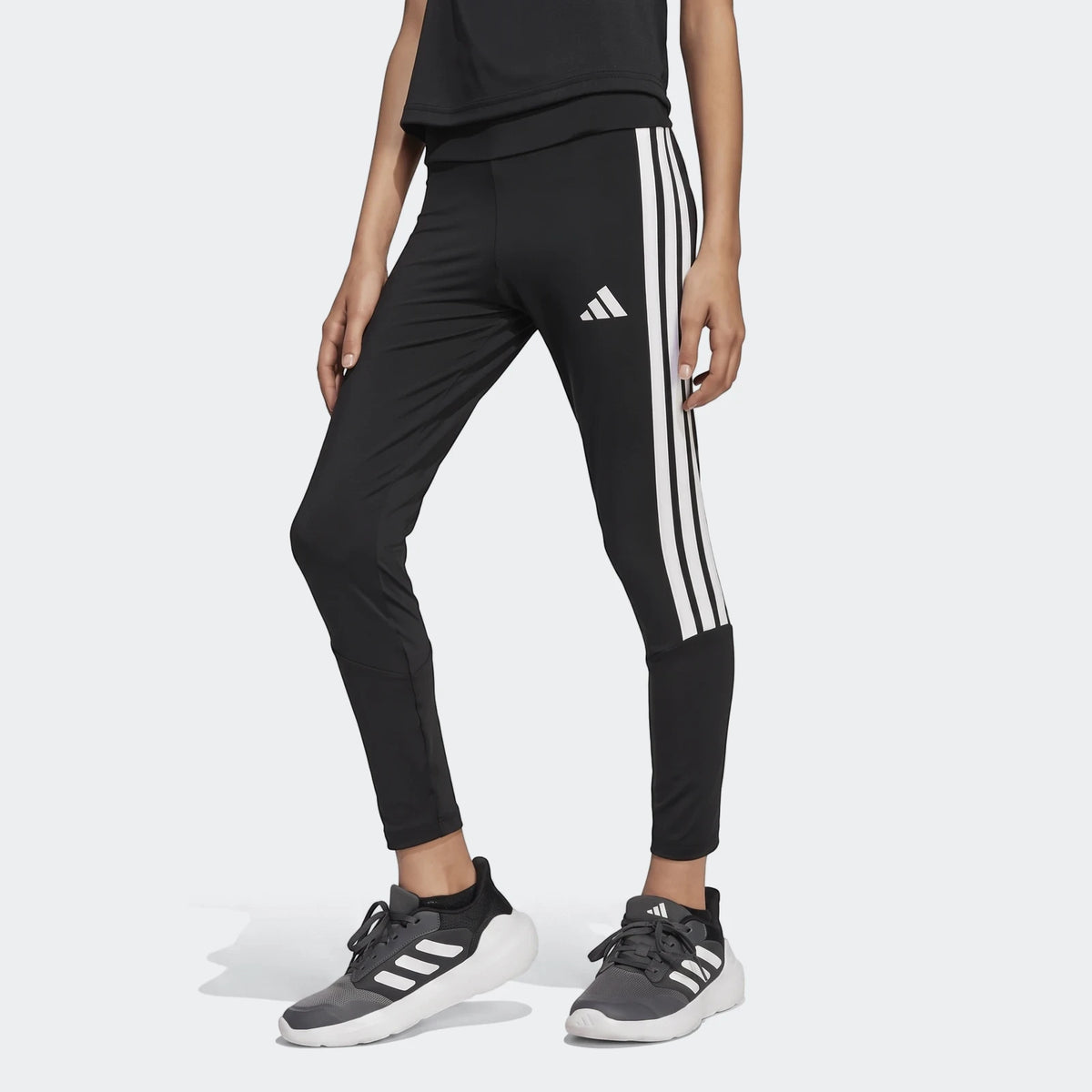 A person models black Adidas Train Essentials 3-Stripes Kids Leggings with white stripes and logo, paired with dark sneakers.