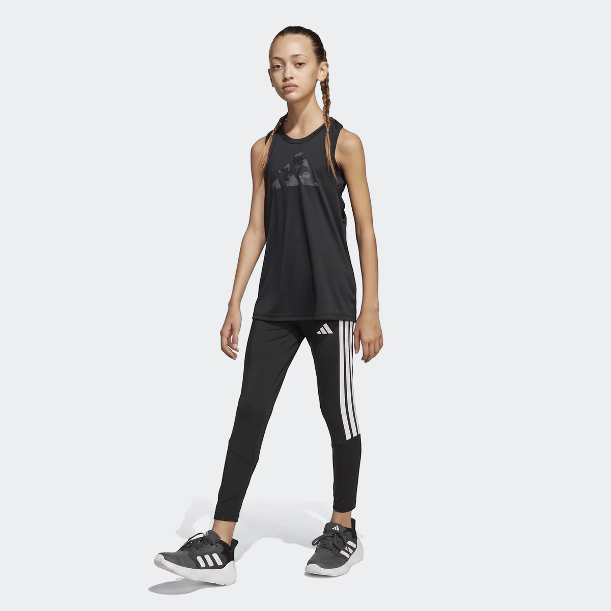 Young girl wearing black Adidas Train Essentials 3-Stripes leggings, a matching tank top, and sneakers for athletic