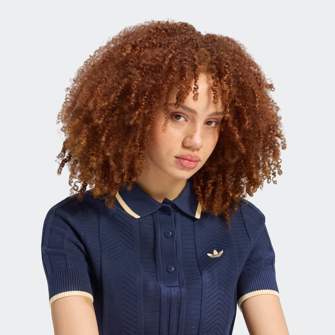 A woman with curly red hair models an Adidas Cable Knit Cali Tee Polo in navy blue with cream stripes on the collar and