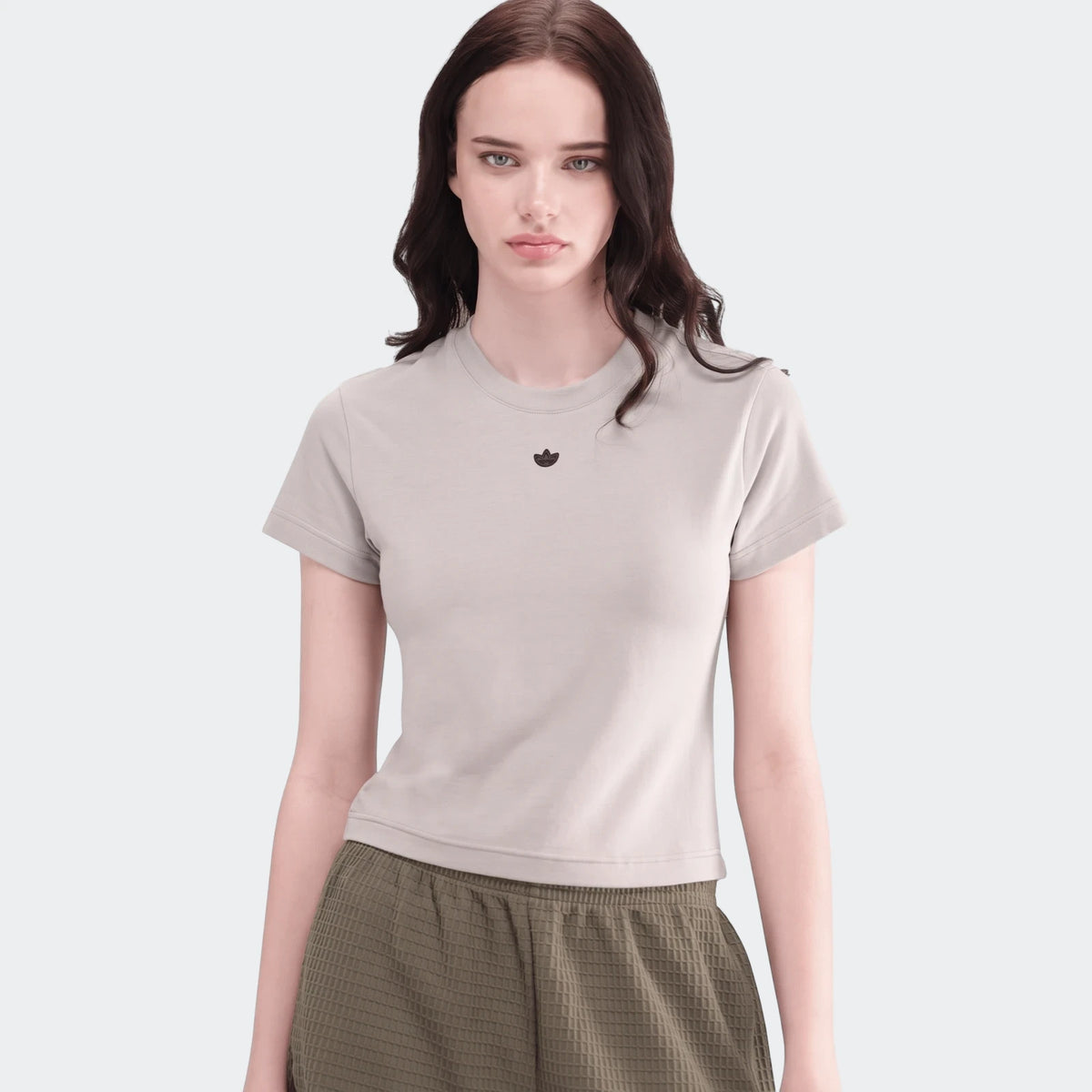 Woman modeling a light beige Adidas t-shirt and olive green textured essential shorts, presented in a casual studio setting.