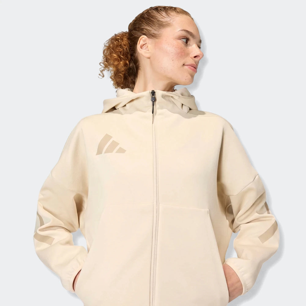 Woman wearing a beige Adidas W Z.N.E. full-zip hoodie, showcasing a relaxed fit and distinct sleeve stripes.