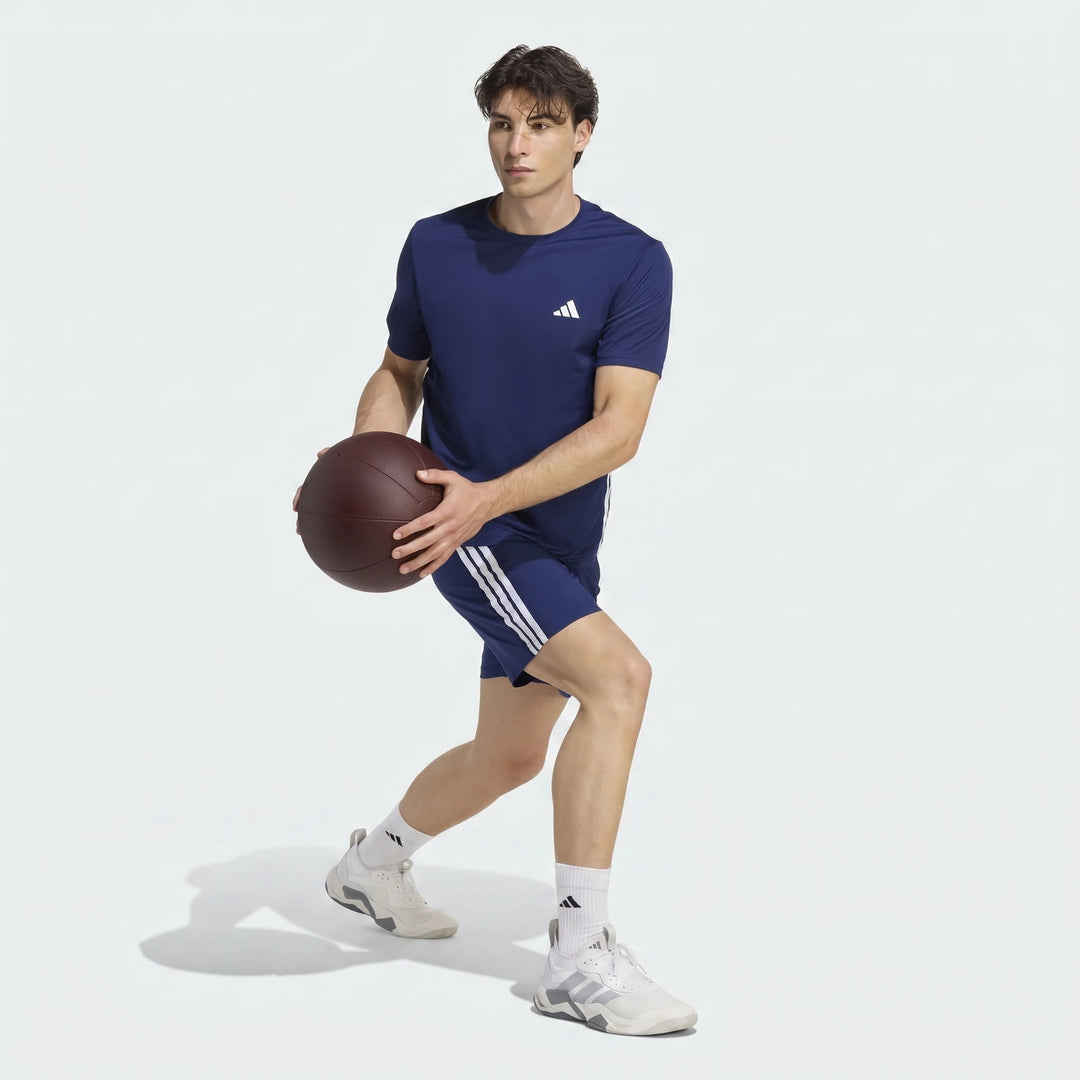Male model wearing navy Adidas Workout Essentials Base 3-Stripes Woven Shorts and t-shirt, holding a medicine ball.