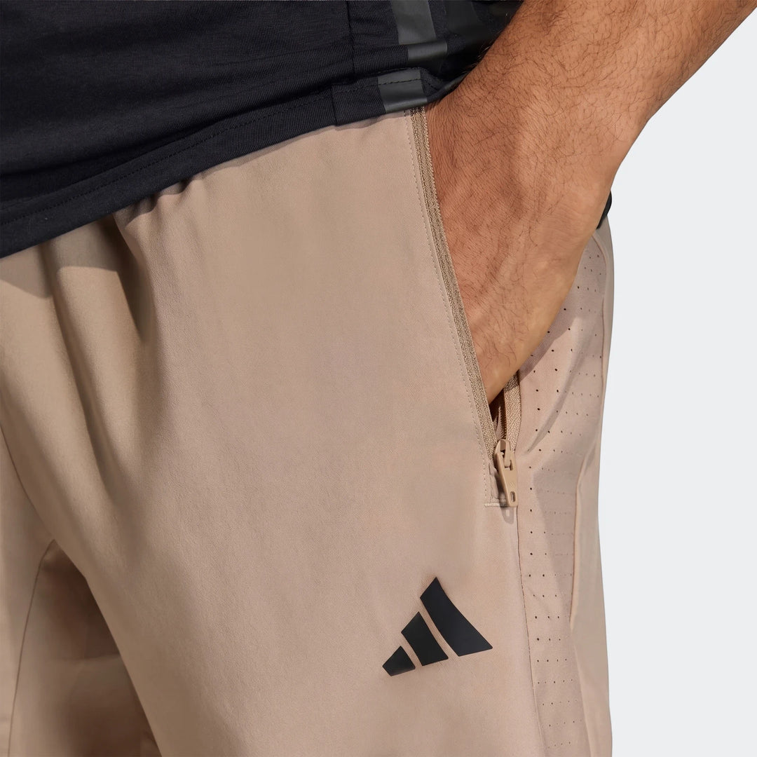 Close-up of beige Adidas Workout Essentials Base Woven Shorts with a black logo and zippered pocket, worn by a person
