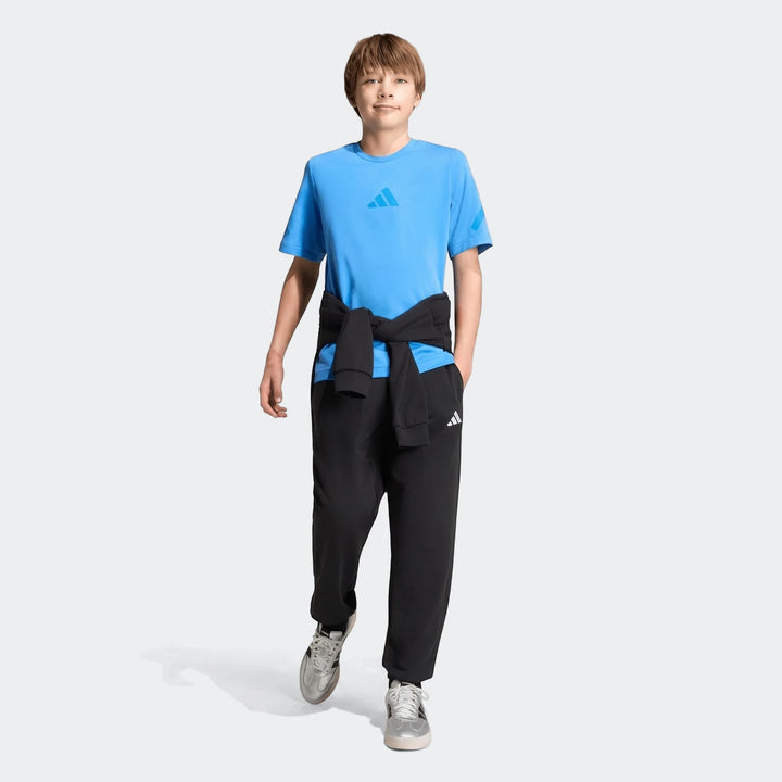 Young boy wearing a blue Adidas Youth Z.N.E. T-shirt, black sweatpants, and silver sneakers in a full body shot.
