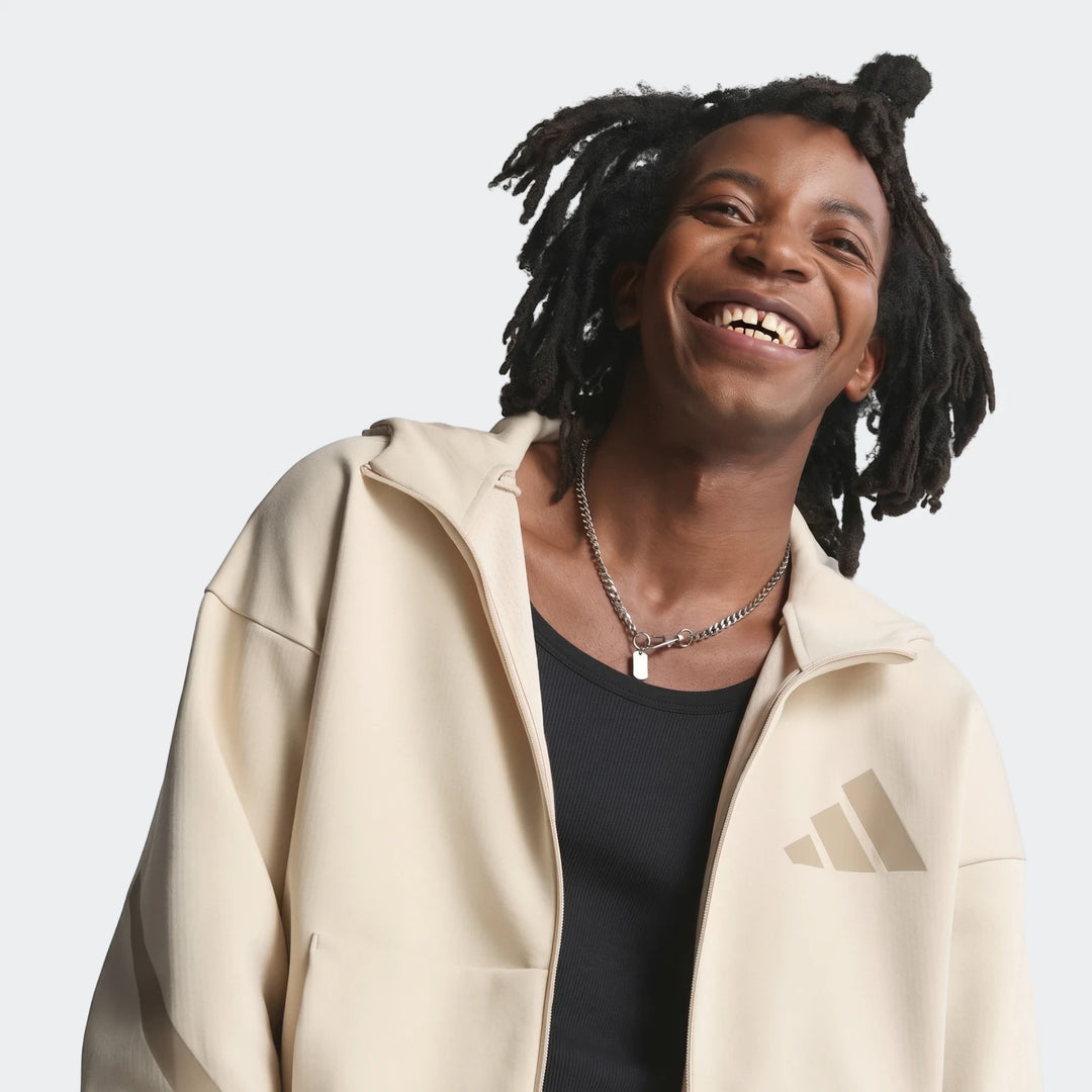 Smiling man with dreadlocks wearing a light beige Adidas Z.N.E. Full-Zip Hoodie and a silver necklace on an off-white