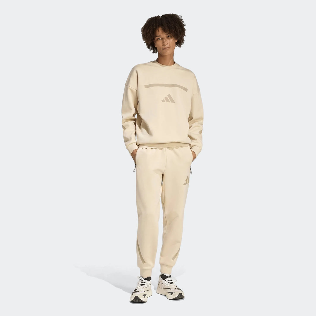 Full body view of a male model wearing beige Adidas Z.N.E. Track Pants and a matching beige sweatshirt, standing with hands