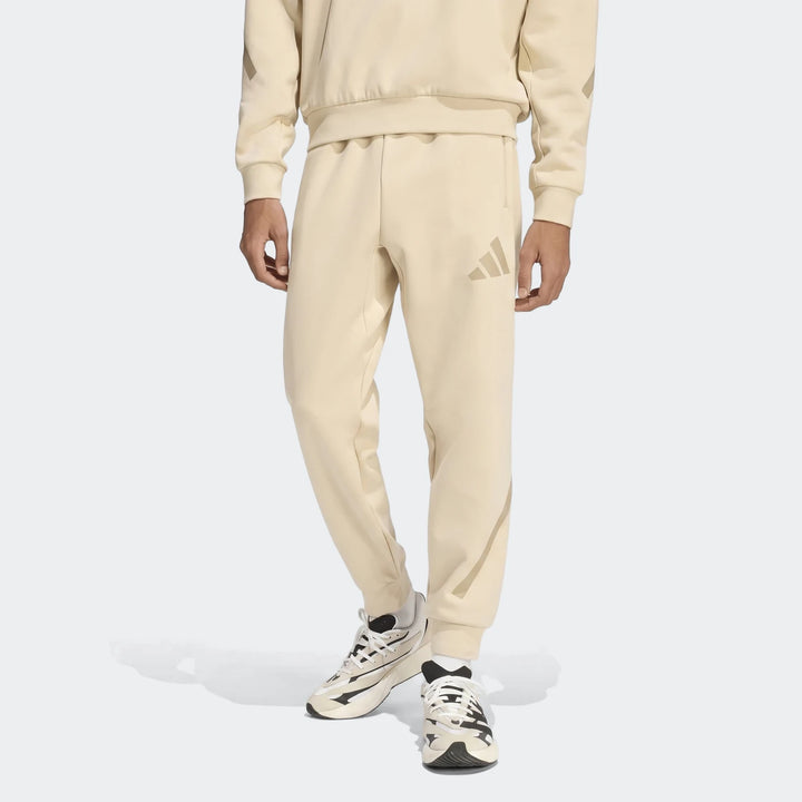 A person wears Adidas Z.N.E. Track Pants in light beige, featuring a subtle logo, paired with black and white sneakers.