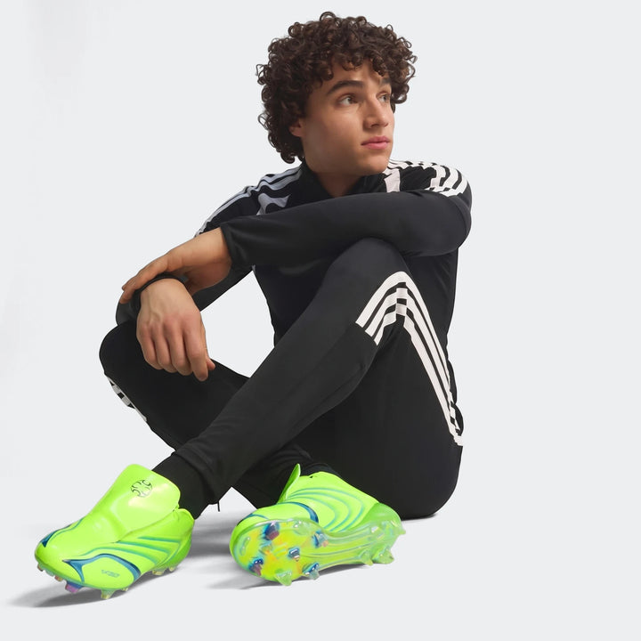 Young man with curly hair wearing a black Adidas Z.N.E. Track Top and track pants, seated with neon green soccer cleats.