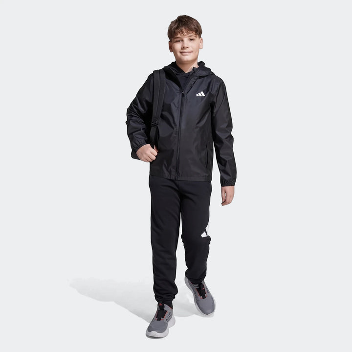 Young boy in a black Adidas Z.N.E. Track Top, black sweatpants, and grey sneakers with a backpack, full body view.