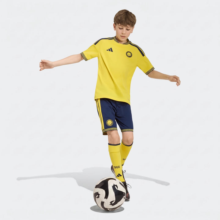 Young boy in yellow and blue Al Nassr FC 26/27 home jersey and shorts balancing a soccer ball.